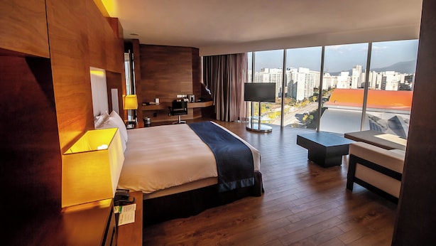 Une chambre d’hôtel moderne dotée d’un grand lit, d’un bureau, d’une chaise et offrant une vue sur la ville grâce à de grandes fenêtres.