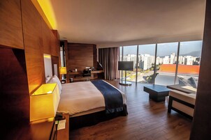 Una habitación de hotel moderna con una cama grande, un escritorio, una silla y vistas a la ciudad a través de amplias ventanas.