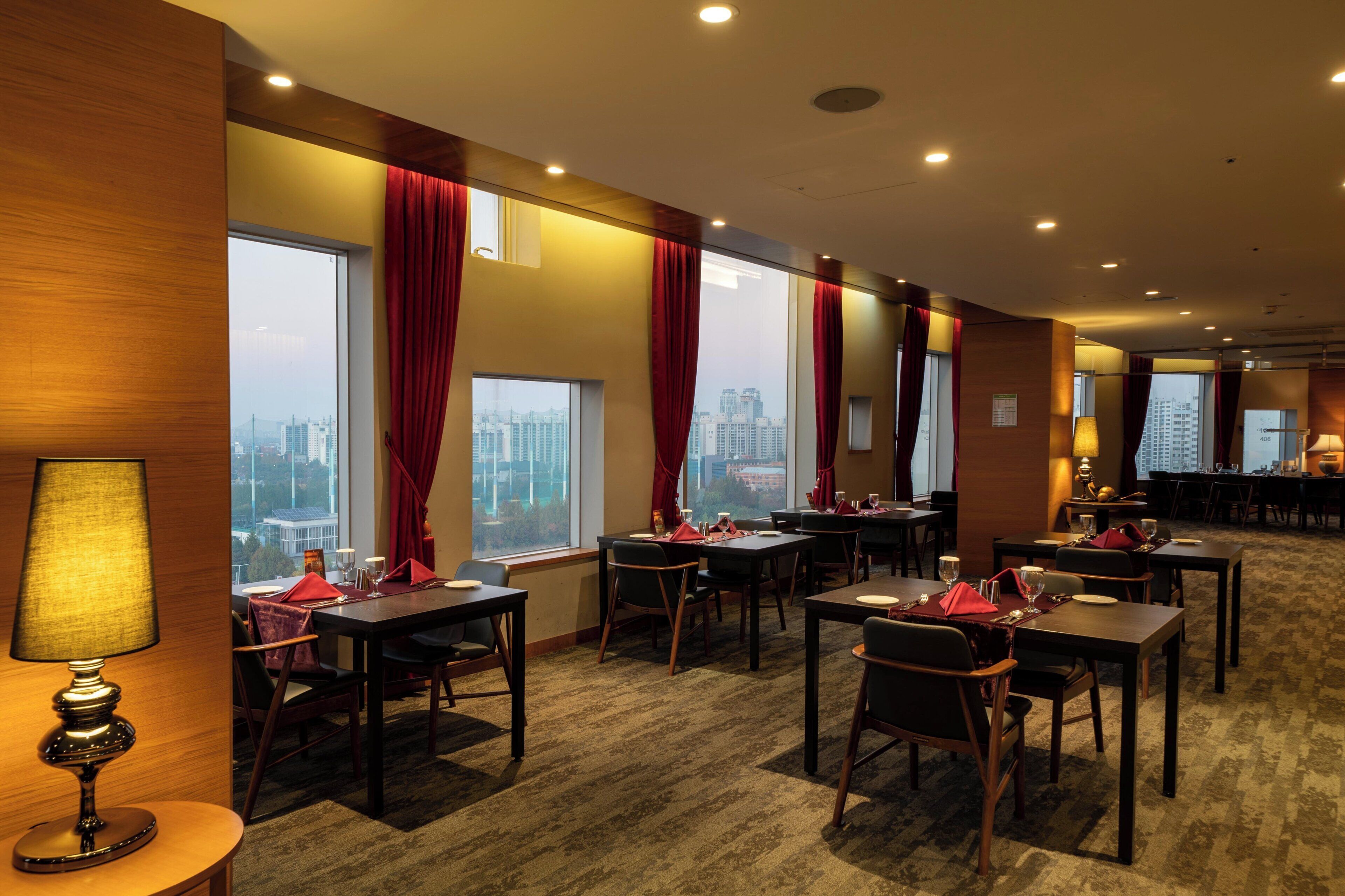 Une salle à manger d’hôtel avec de nombreuses tables et chaises, de grandes fenêtres avec des rideaux et une vue sur la ville.