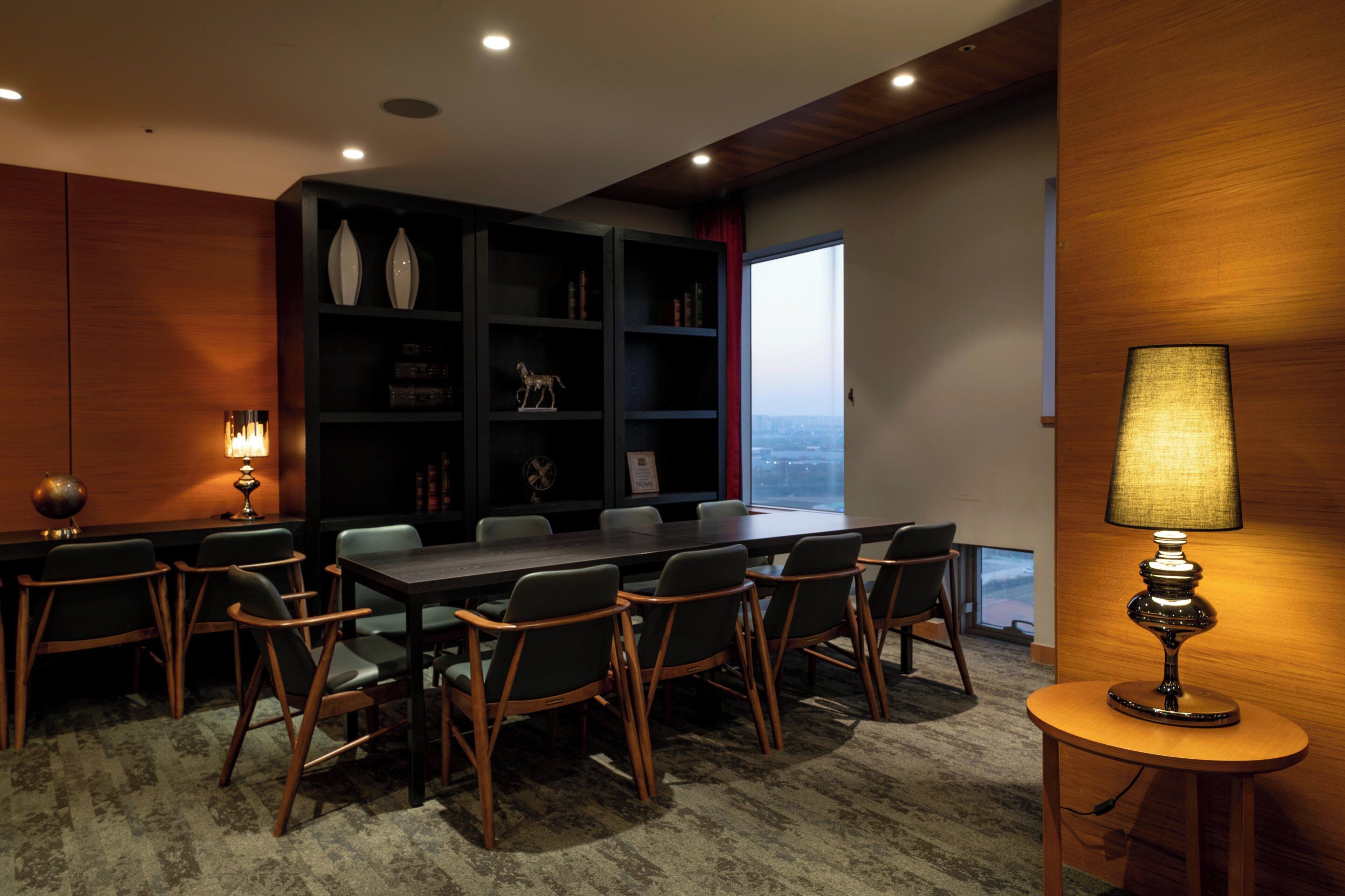 Un salón de conferencias con una mesa de madera oscura, sillas, una estantería y una lámpara.