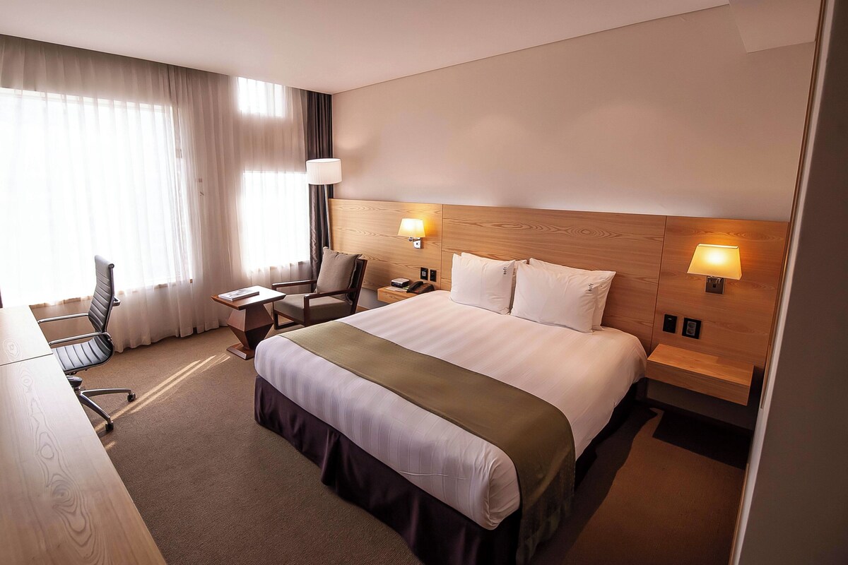 Habitación de hotel con una cama grande, dos mesitas de noche con lámparas, un escritorio con una silla y una ventana con cortinas.
