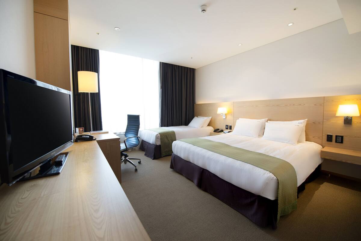 A hotel room with two beds, a desk with a TV, a chair, and a large window with curtains.