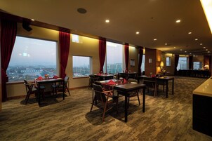 Un comedor de hotel con varias mesas y sillas, ventanas amplias con cortinas y suelo alfombrado.