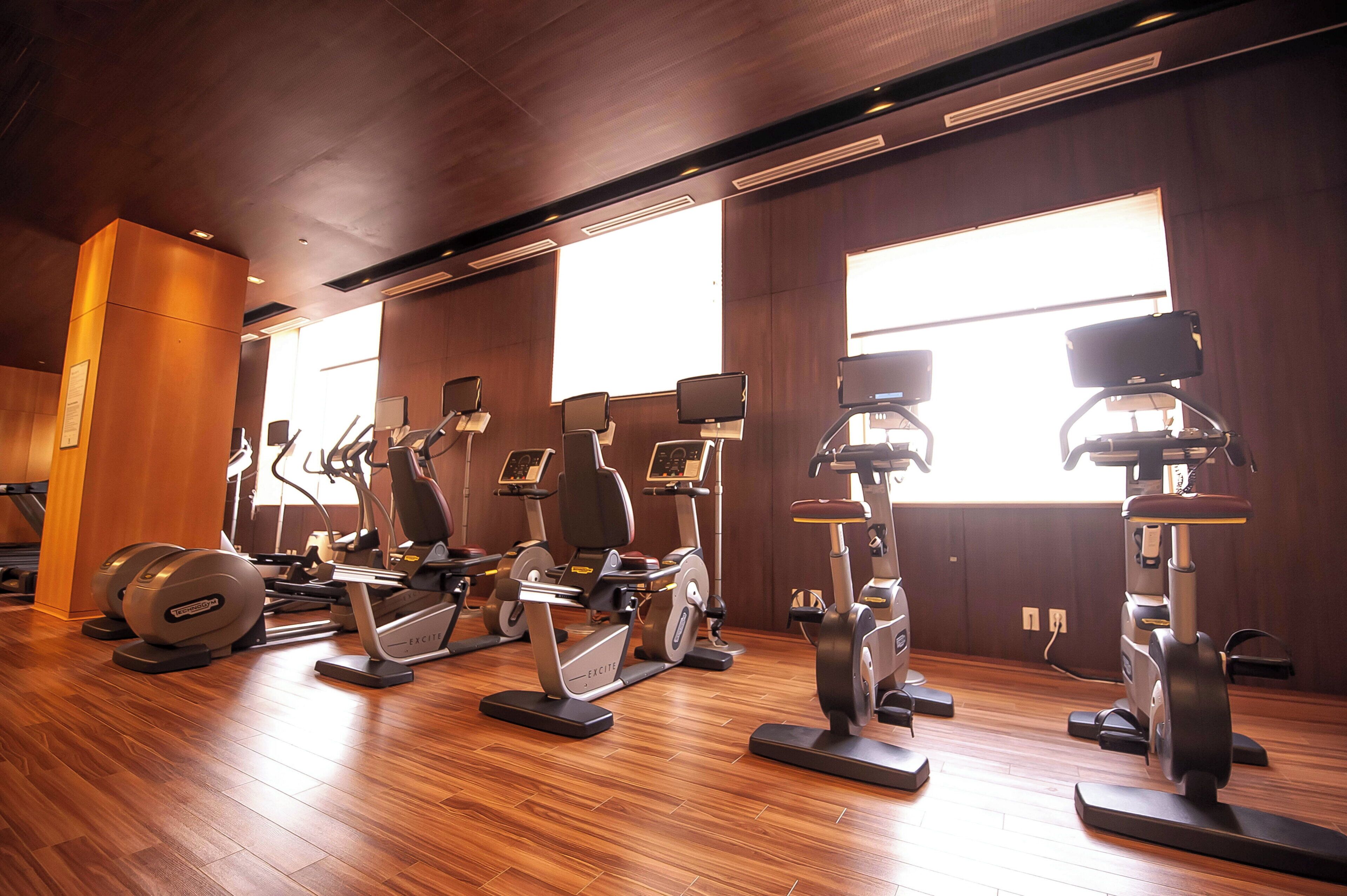 Un gimnasio con diversas máquinas de ejercicio, suelo de madera y amplios ventanales.