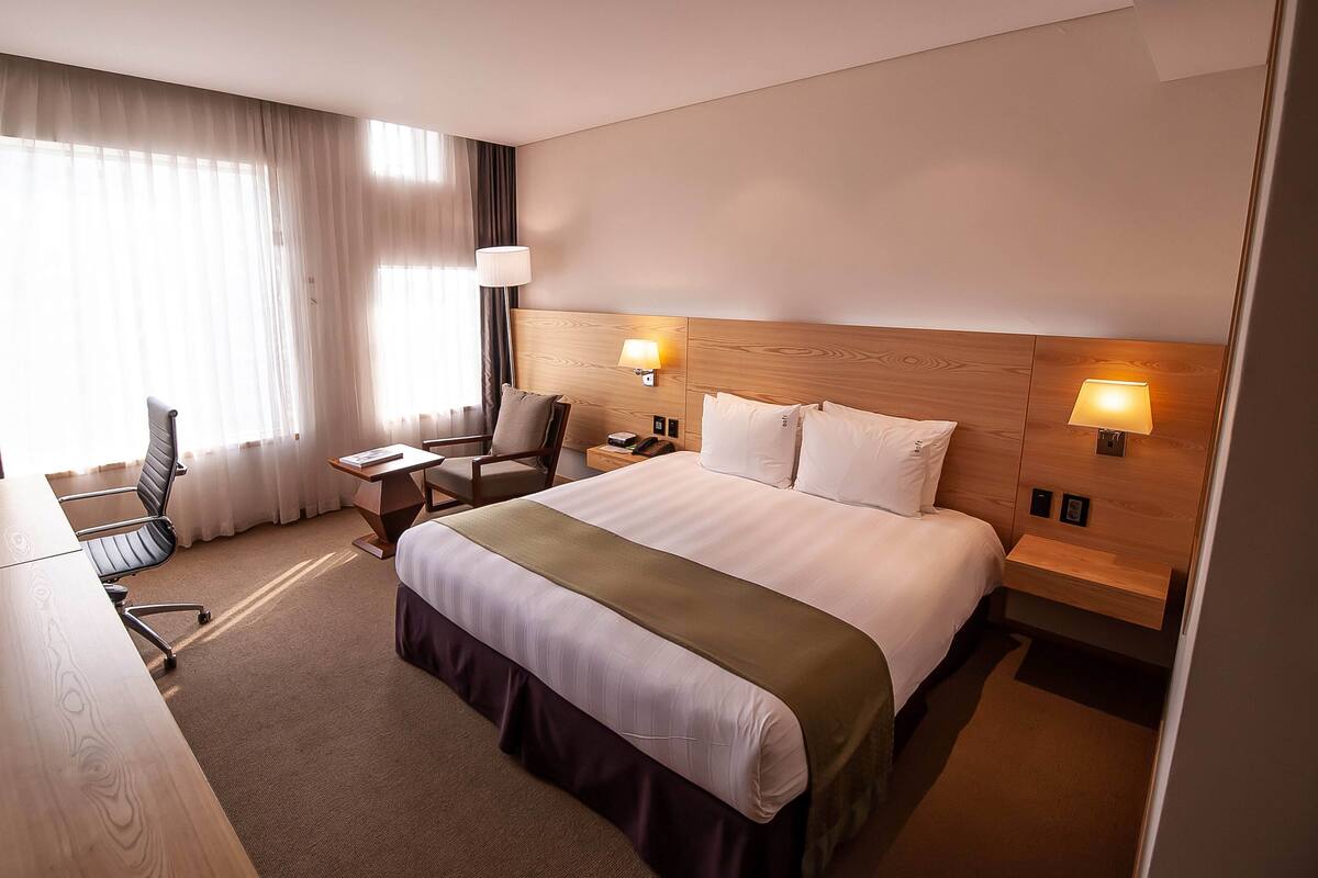 A hotel room with a large bed, two bedside lamps, a desk with a chair, and a window with curtains.