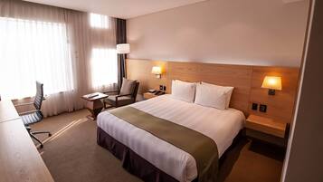 A hotel room with a large bed, two bedside lamps, a desk with a chair, and a window with curtains.