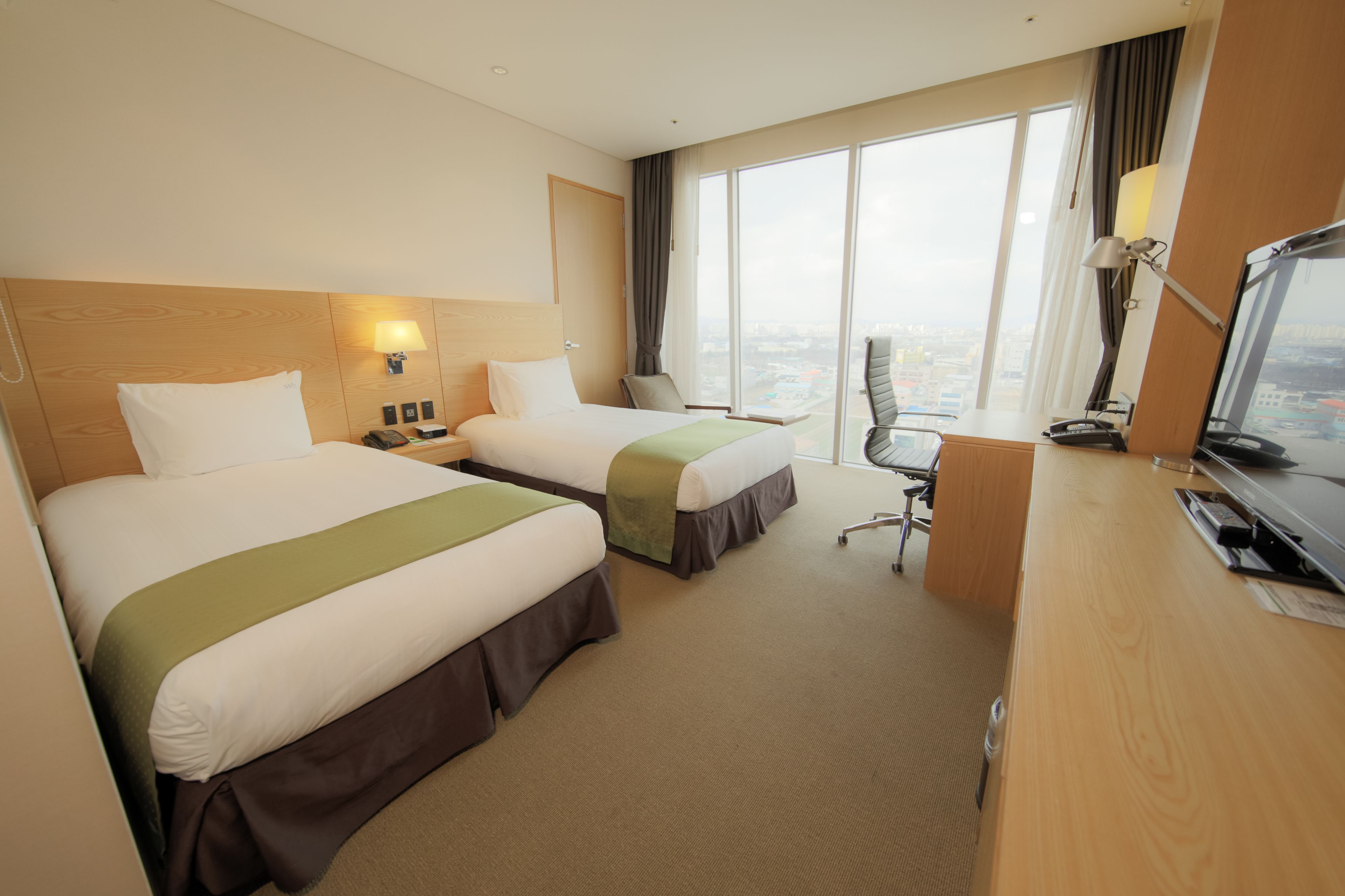 A hotel room with two beds, a desk, a chair, and a large window offering a city view.