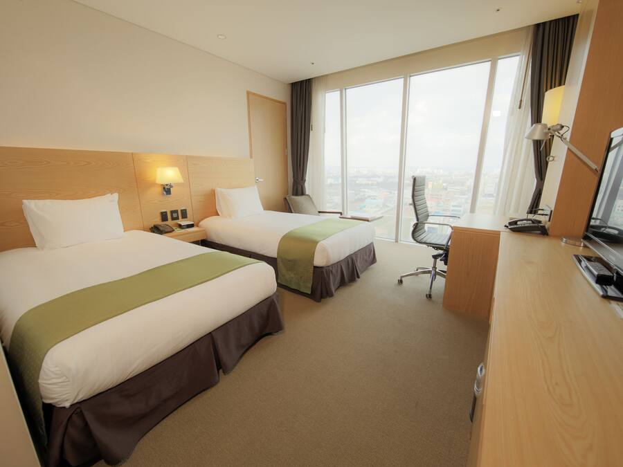 A hotel room with two beds, a desk, a chair, and a large window offering a city view.