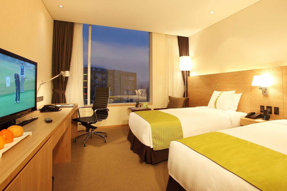 A hotel room with two beds, a desk with a TV, a chair, and a view of the city at dusk.