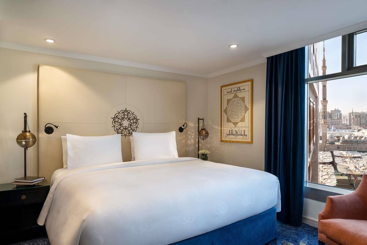 Een hotelkamer met een groot bed, uitzicht op een stadsbeeld, een citaat in een lijst op de muur en een nachtkastje met een lamp.