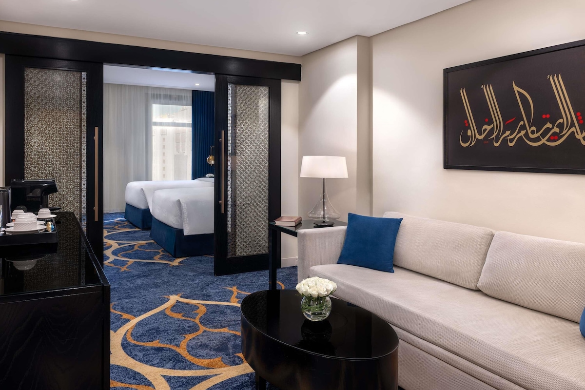 Habitación de hotel con cama, sofá, mesa de centro, escritorio con lámpara y un adorno en la pared.