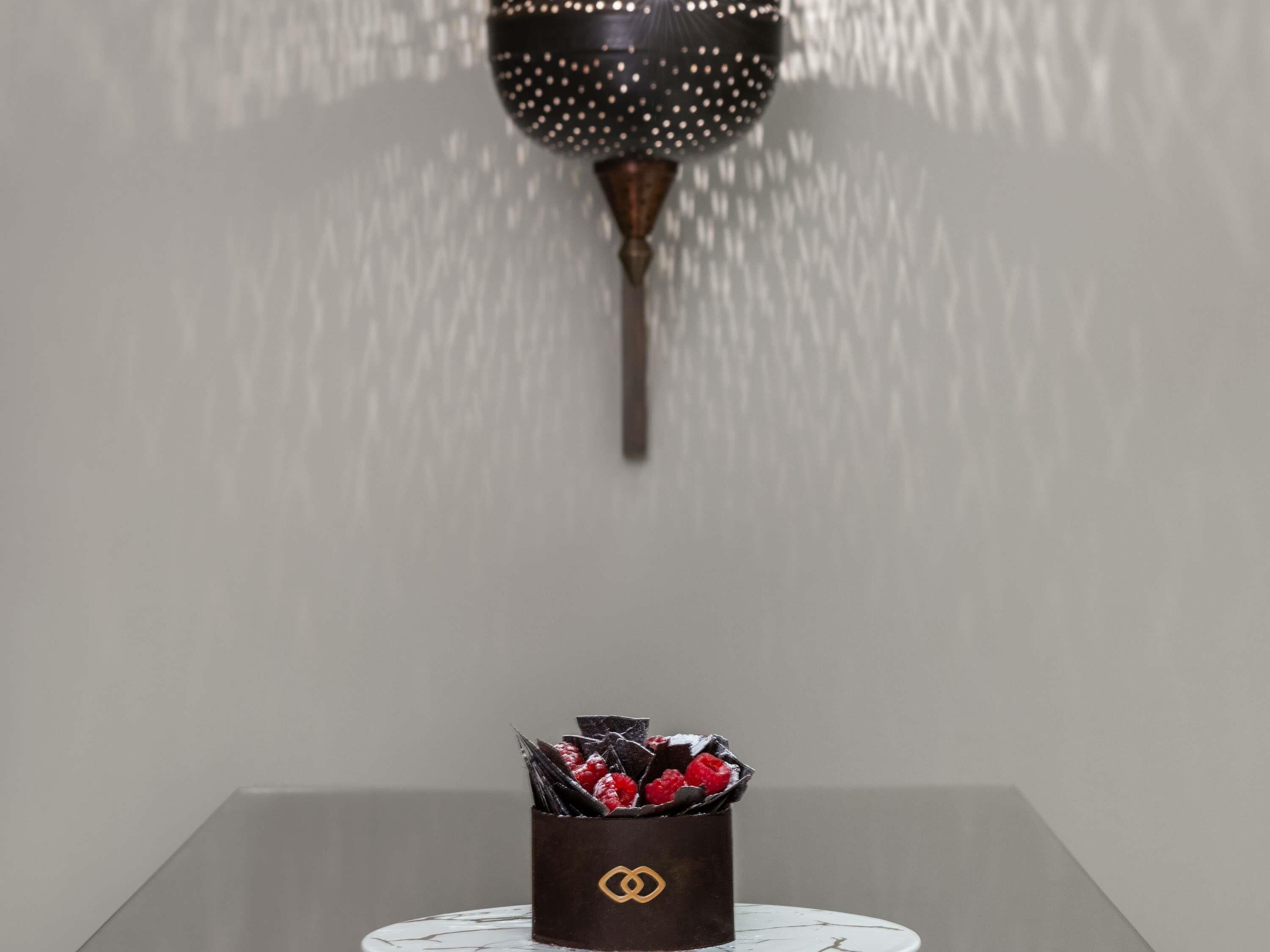 A table with a chocolate dessert topped with raspberries and chocolate shavings, a white plate with a gold logo, and a wall-mounted light fixture.