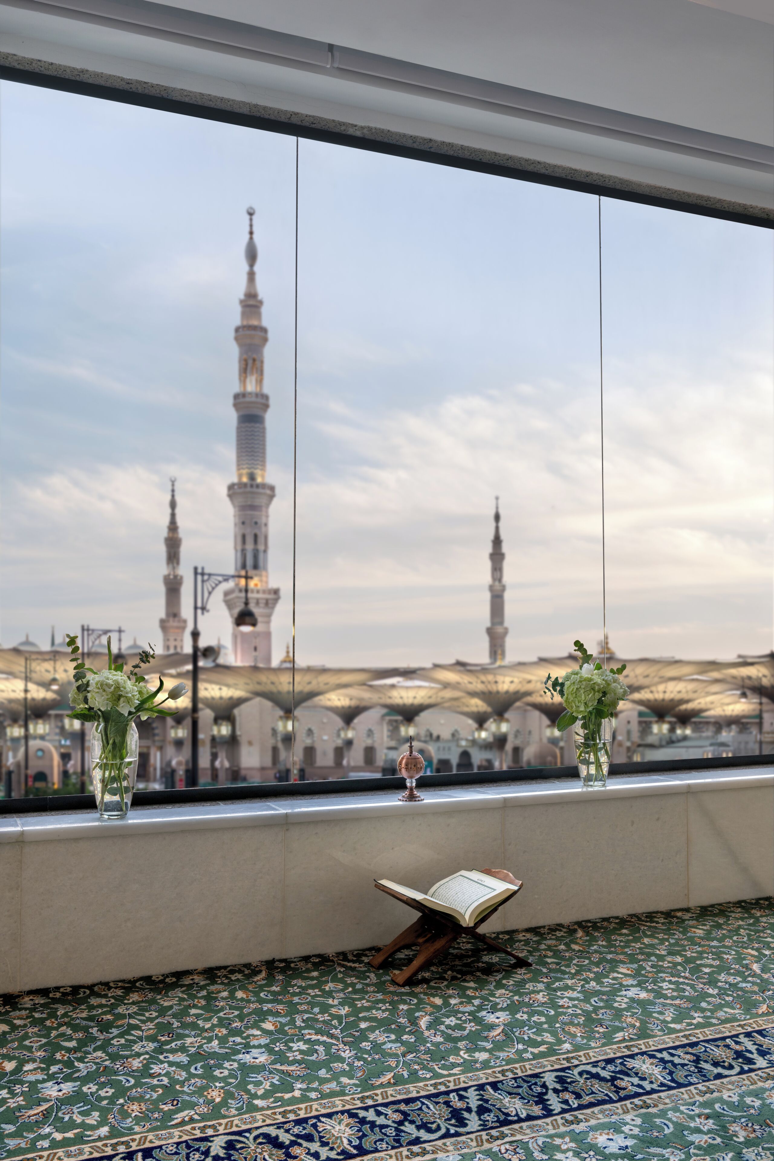 Une vue sur une mosquée avec ses minarets, aperçue à travers une fenêtre, un tapis à motifs et une petite bibliothèque.