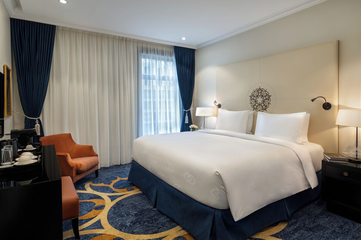 A hotel room with a large bed, a desk with a chair, a nightstand, and a wall-mounted lamp.