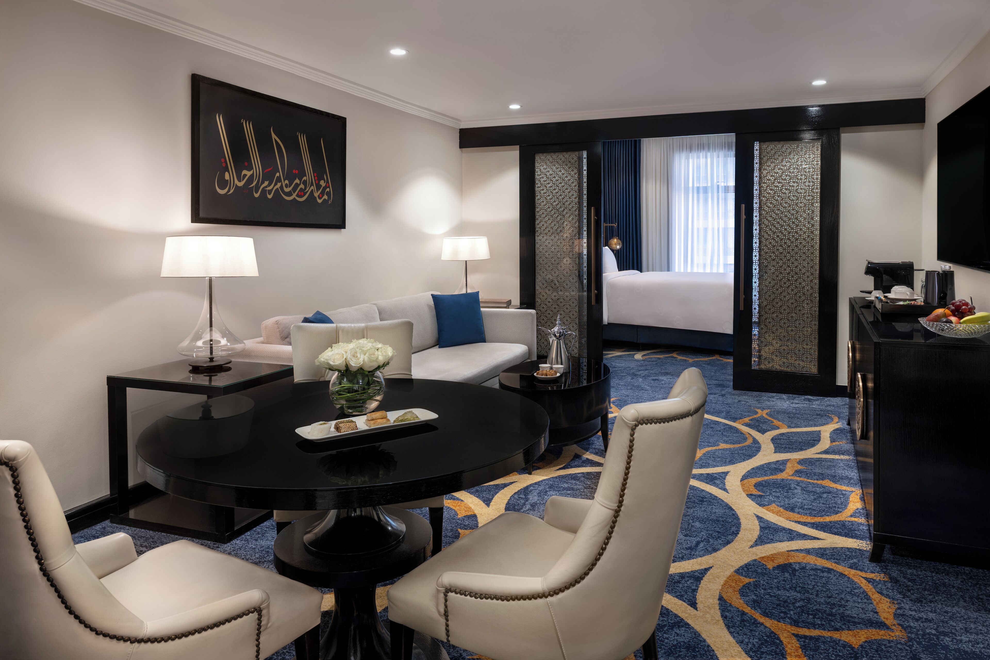 A hotel room with a dining table, two chairs, a sofa, a bed, a TV, and a decorative wall piece.