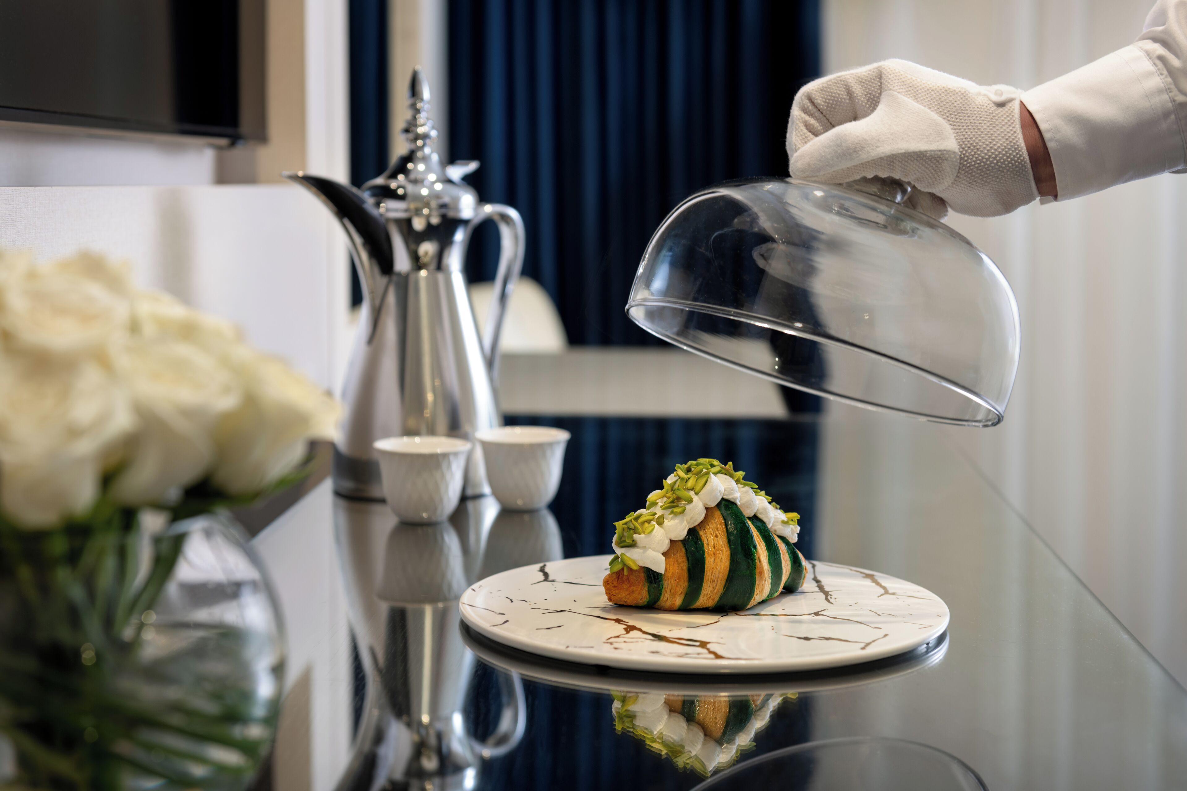 Un dessert présenté sur une assiette, recouvert d’un couvercle en forme de dôme transparent, une carafe en argent et un vase rempli de fleurs blanches, le tout posé sur une surface réfléchissante.