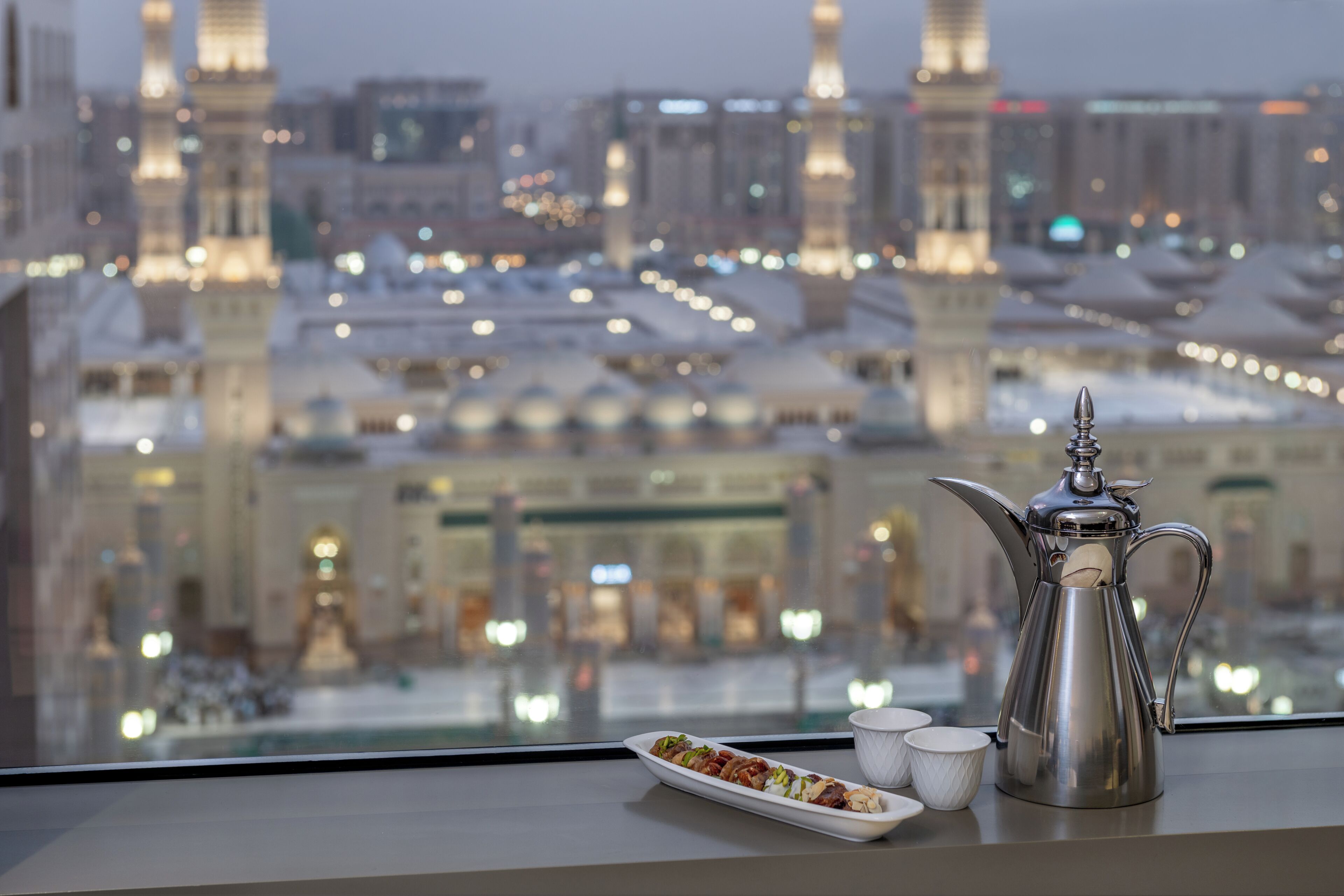 A cityscape with a view of a mosque and buildings, a silver coffee pot, a plate with food, and three cups on a table.