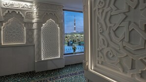 A room with intricate white wall carvings, a patterned carpet, and a view of a tall tower through a window. - Sofitel Shahd Al Madinah (Madinah)