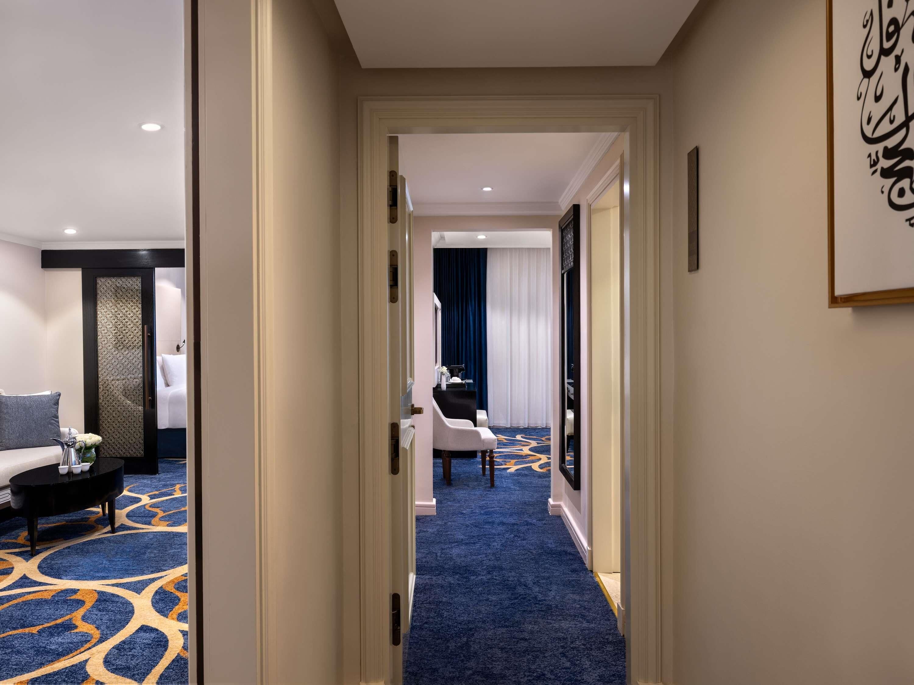 A hotel hallway with a blue carpet, a bed with white linens, a black coffee table, and a decorative wall piece.