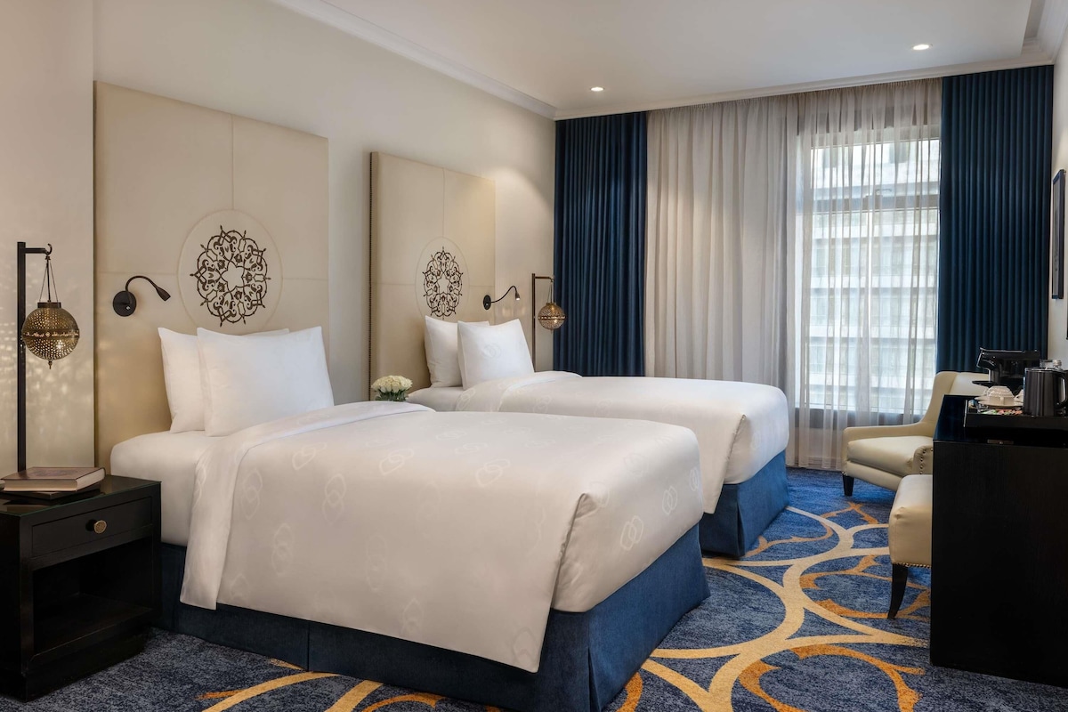Habitación de hotel con dos camas, un escritorio, una silla y un gran ventanal con cortinas.