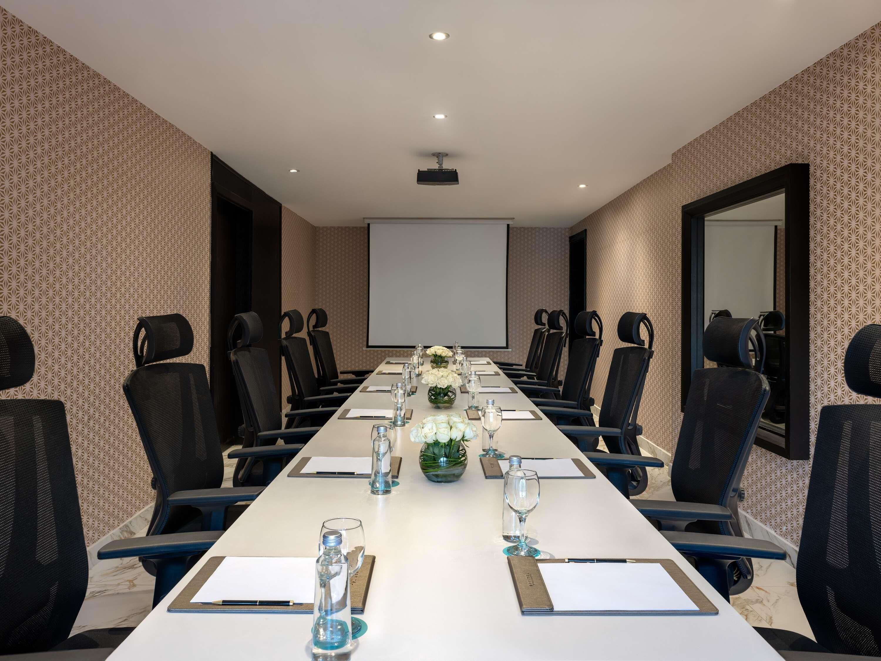 A conference room with a long table, black chairs, a projector screen, and floral centerpieces.