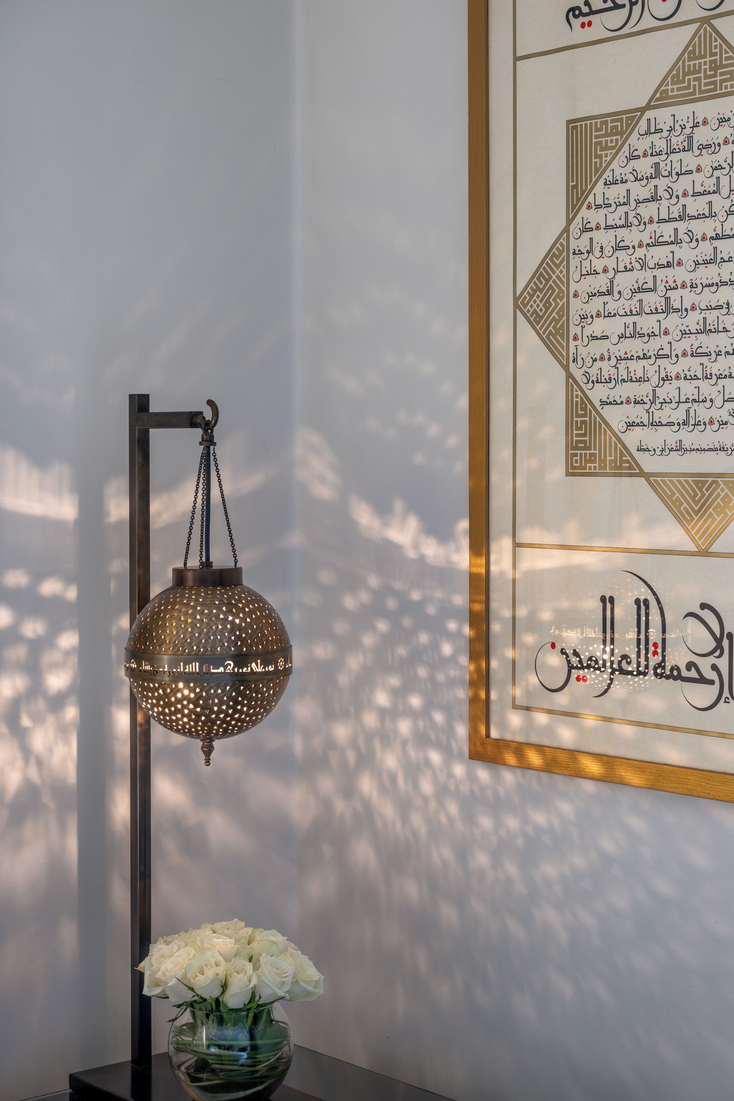 A framed Arabic calligraphy on a wall, a hanging lantern, and a vase with white flowers on a table.