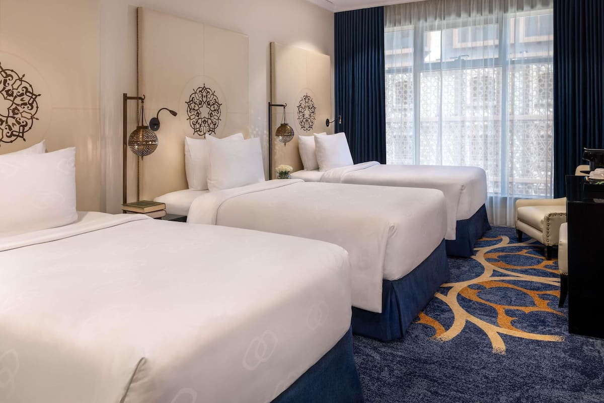 Een hotelkamer met drie bedden, een bureau, een stoel en een groot raam met uitzicht.