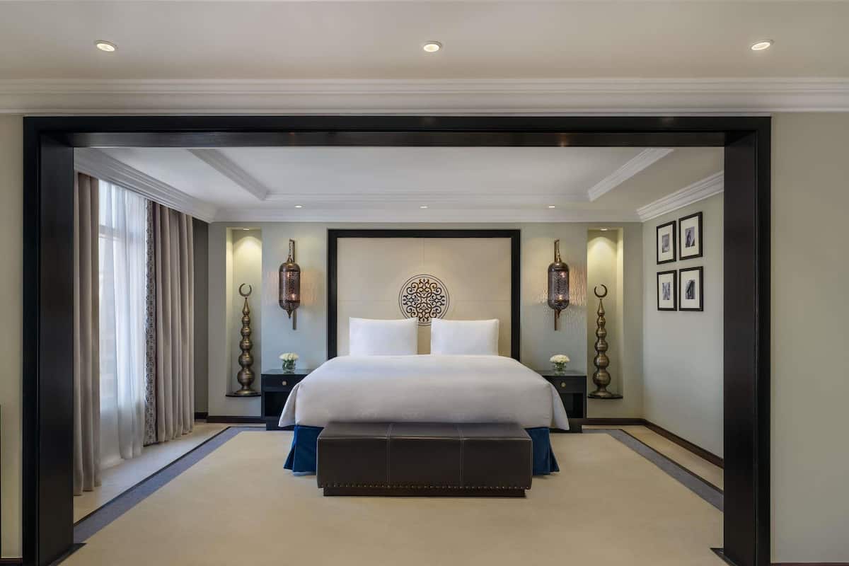 Hotelkamer met een groot bed, twee nachtkastjes, een decoratief wandobject en uitzicht door een geopende deur.