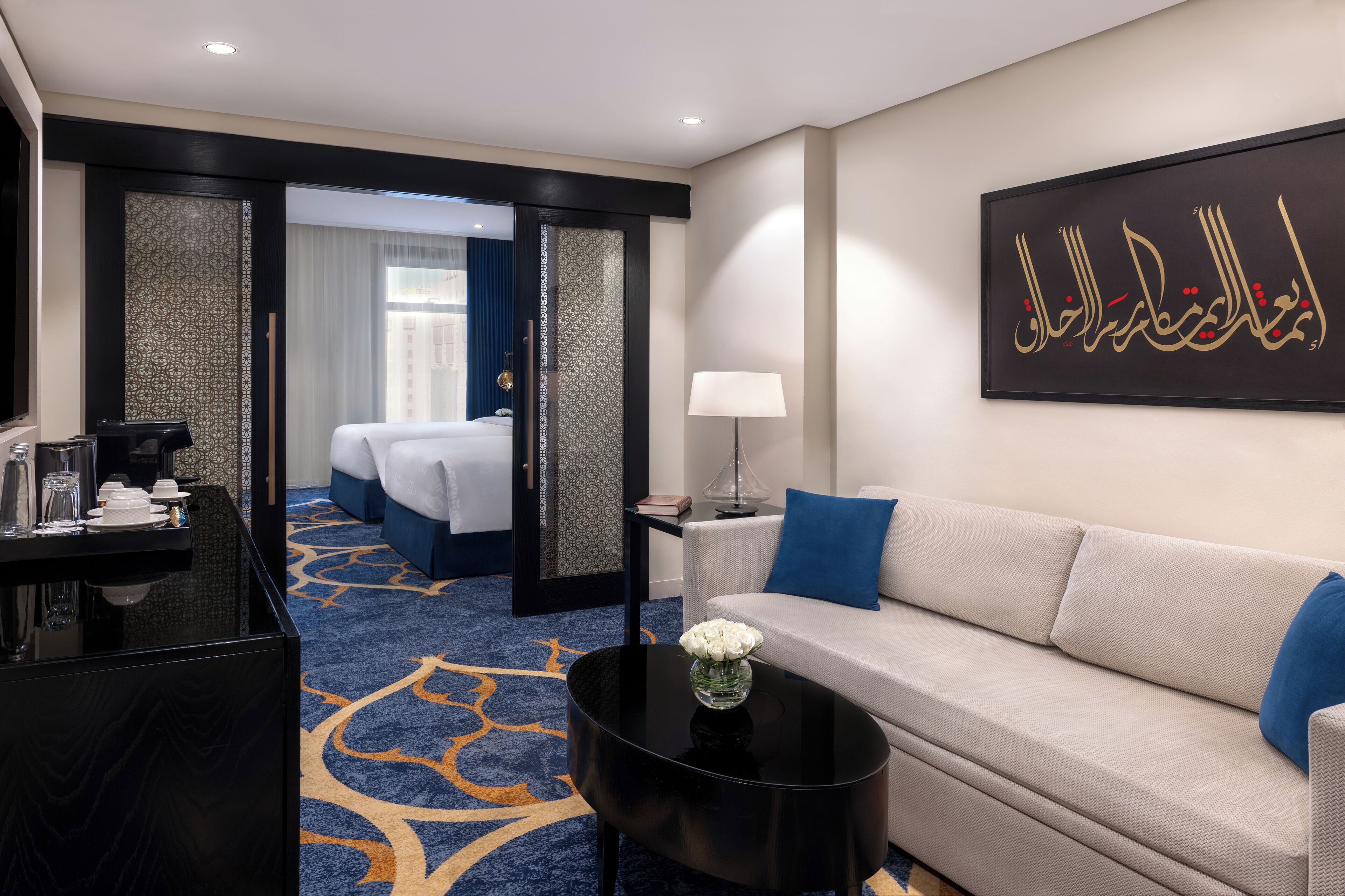 A hotel room with a bed, a sofa, a coffee table, a desk, and a wall with Arabic calligraphy.
