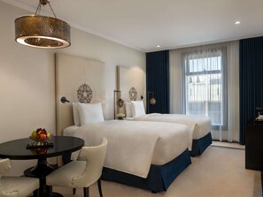 Une chambre d’hôtel avec deux lits, une table à manger, des chaises, un lustre et une grande fenêtre avec des rideaux.