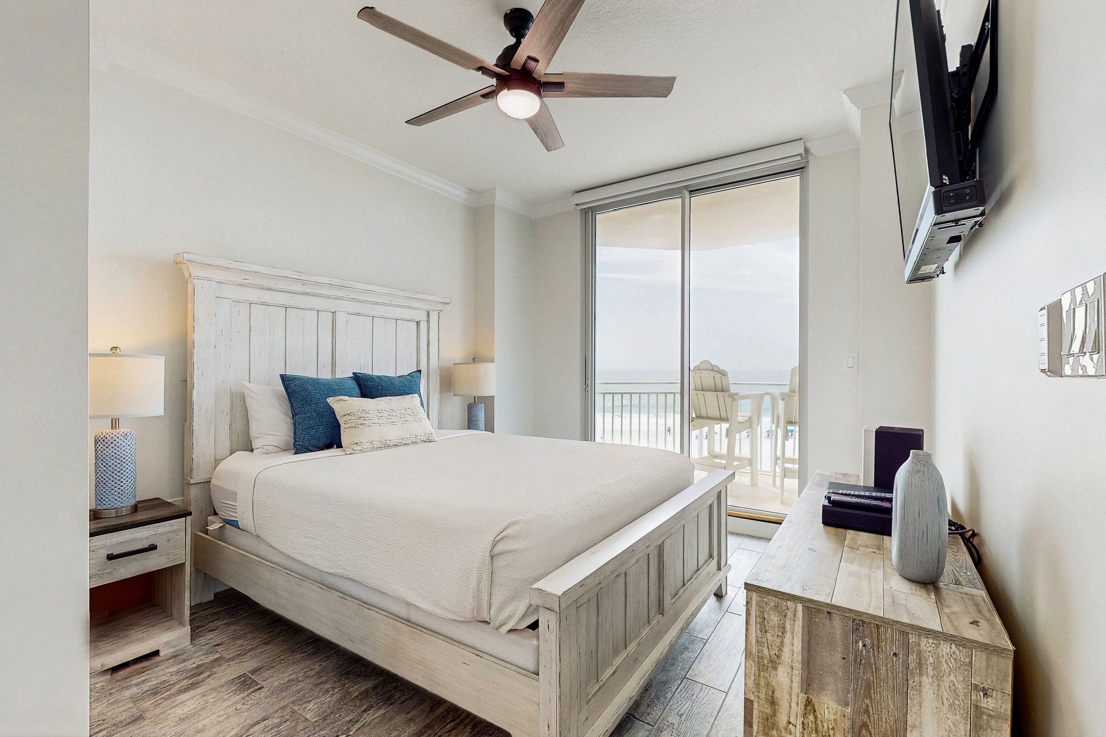A bedroom with a wooden bed, a ceiling fan, a TV mounted on the wall, and a balcony with a view.