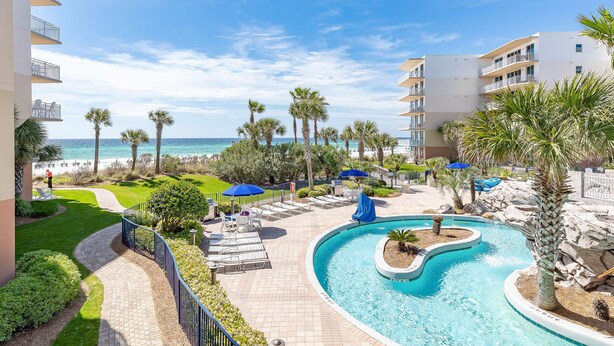 A resort pool area with a curved pool, lounge chairs, and palm trees. - Waterscape Condominiums (Fort Walton Beach)
