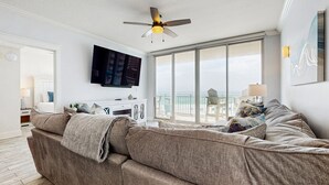 A modern living room with a large gray sectional sofa, a ceiling fan, a flat-screen TV, and a large window offering a view of the outdoors. - Waterscape Condominiums (Fort Walton Beach)
