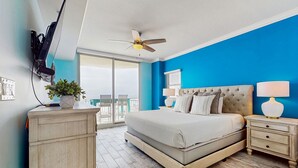 A bedroom with a large bed, a ceiling fan, a nightstand with a lamp, a TV mounted on the wall, and a sliding glass door leading to a balcony. - Waterscape Condominiums (Fort Walton Beach)