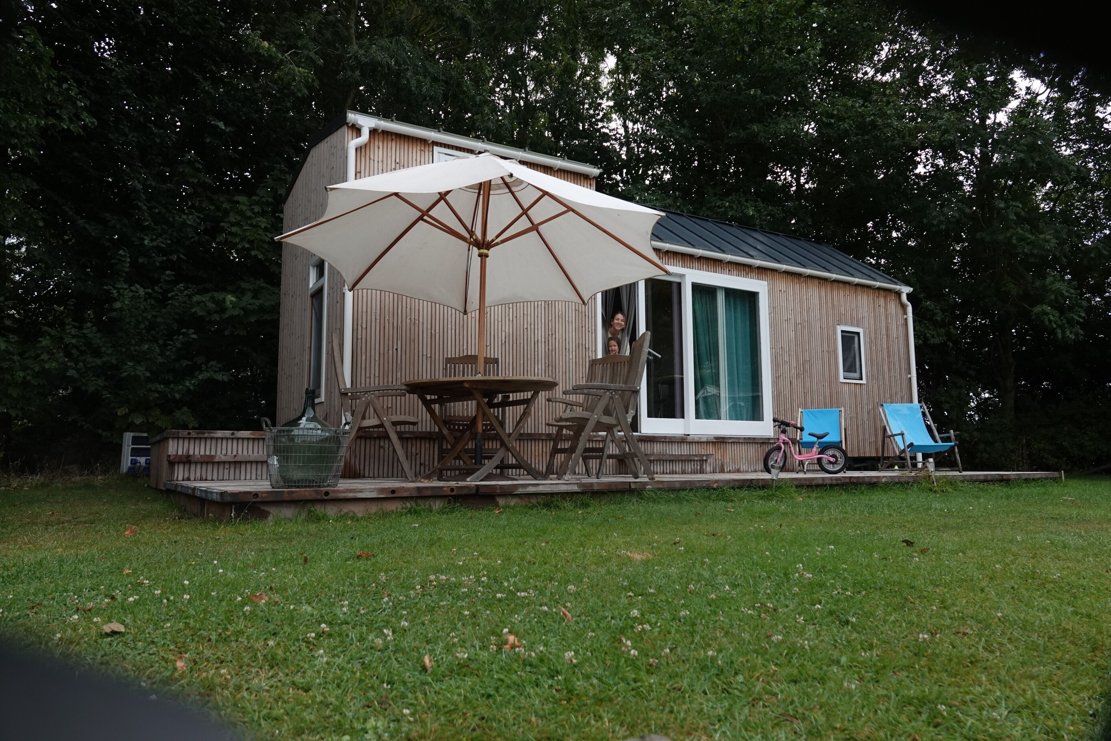 En træhytte med terrasse, et bord, stole, en cykel og en stor parasol.