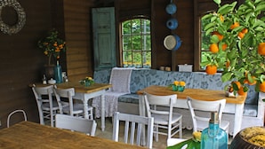 Un comedor rústico con mesas de madera y sillas blancas, un jarrón azul y una planta en maceta con naranjas.