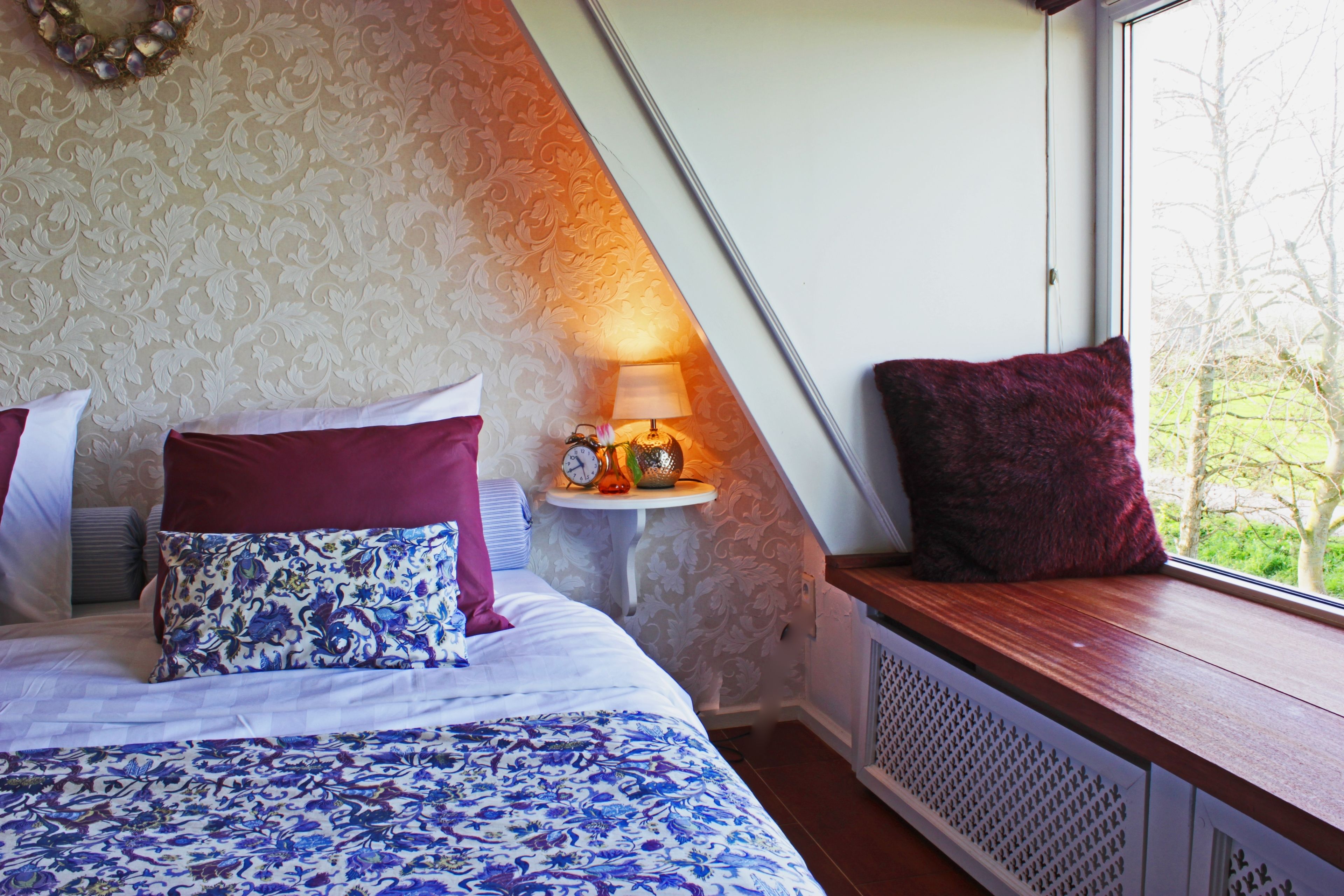 A bedroom with a bed, pillows, a bedside table with a lamp, and a window with a view of trees.