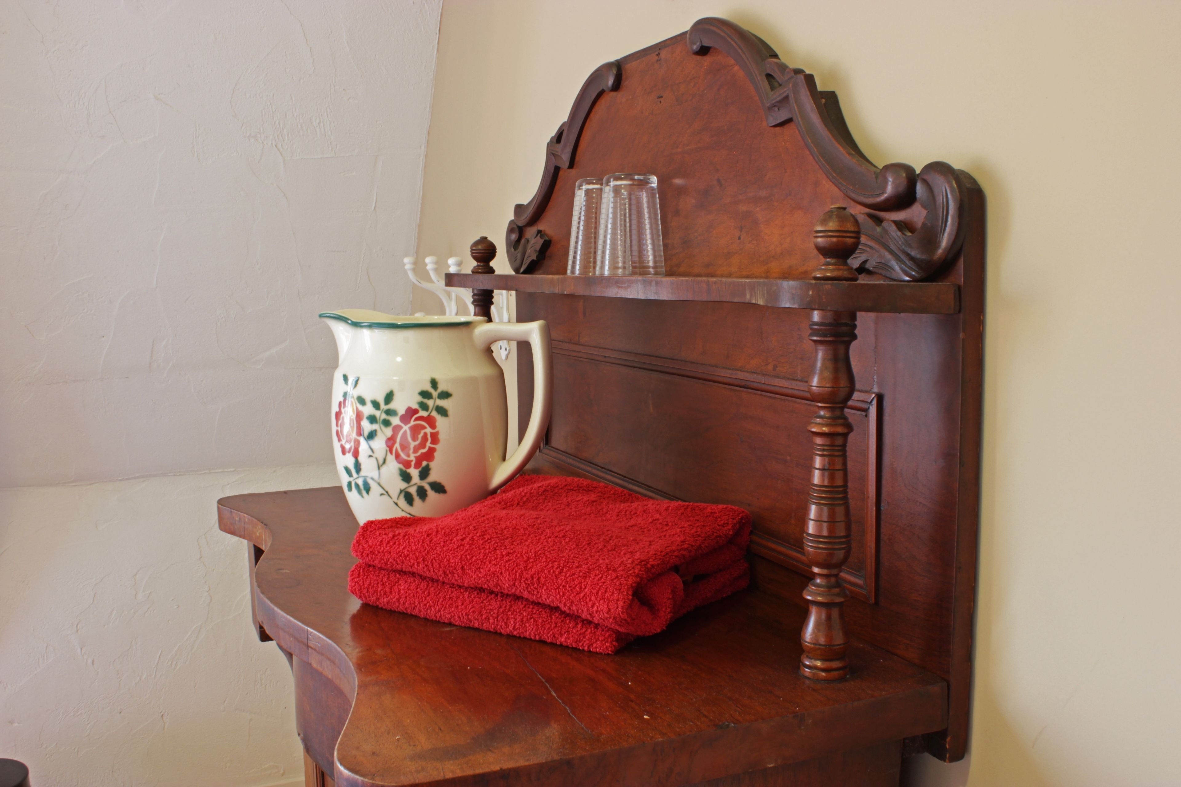 A wooden nightstand with a white pitcher featuring floral designs, a red towel, and two clear glasses.