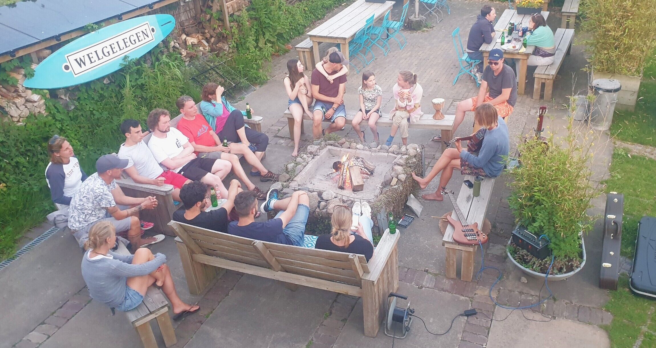 Un grupo de personas sentadas alrededor de un brasero en un entorno al aire libre, con un banco de madera, mesas y sillas.