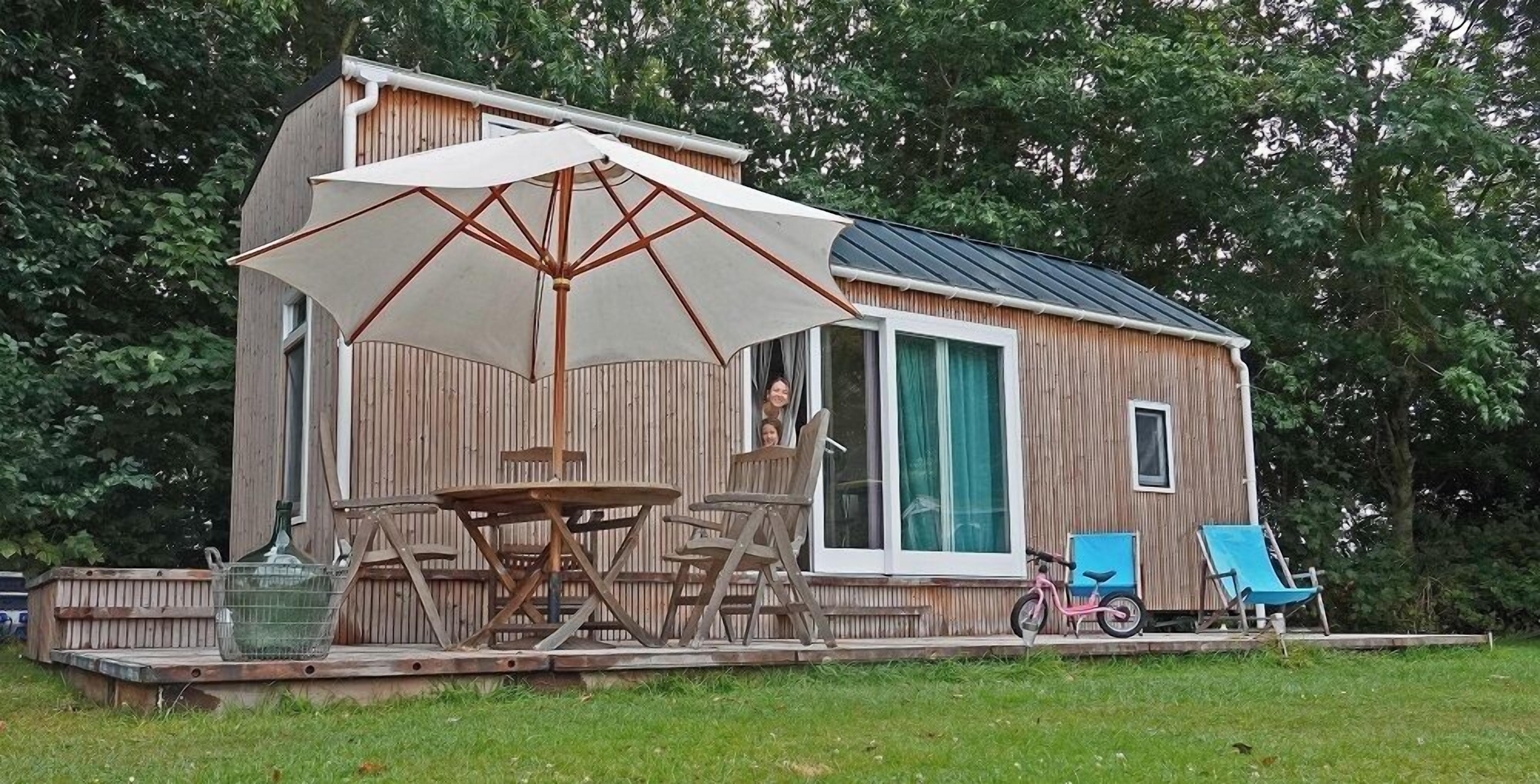 A wooden cabin with a deck, a table with chairs, a large umbrella, and a bicycle.