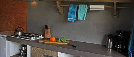 Une cuisine équipée d’une cuisinière, d’une planche à découper et de divers ustensiles de cuisine. Des étagères au-dessus du plan de travail servent à ranger les assiettes et les tasses.
