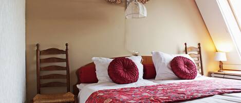 A bedroom with a wooden chair, a bed with red and white bedding, two wall-mounted wreaths, and a nightstand with a lamp.