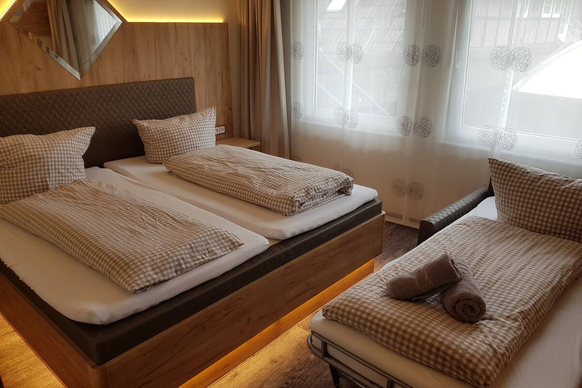 A double bed with checkered bedding and a wooden headboard, a bedside table, and a window with sheer curtains.