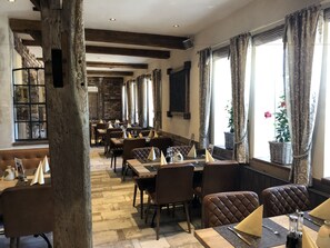 Un restaurante con vigas de madera, paredes de piedra y ventanas amplias con cortinas. Las mesas están dispuestas con servilletas, cubiertos y saleros y pimenteros.