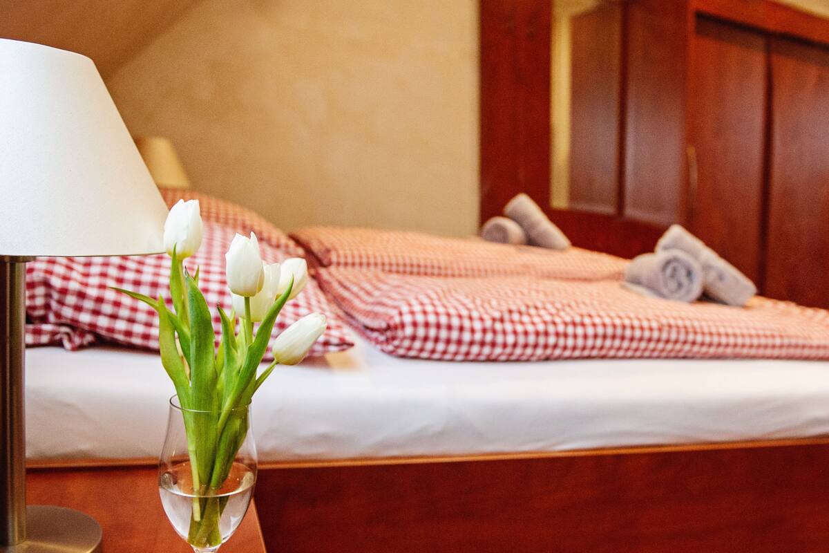 A bed with a checkered blanket and pillows, a bedside table with a lamp, and a glass vase with white flowers.