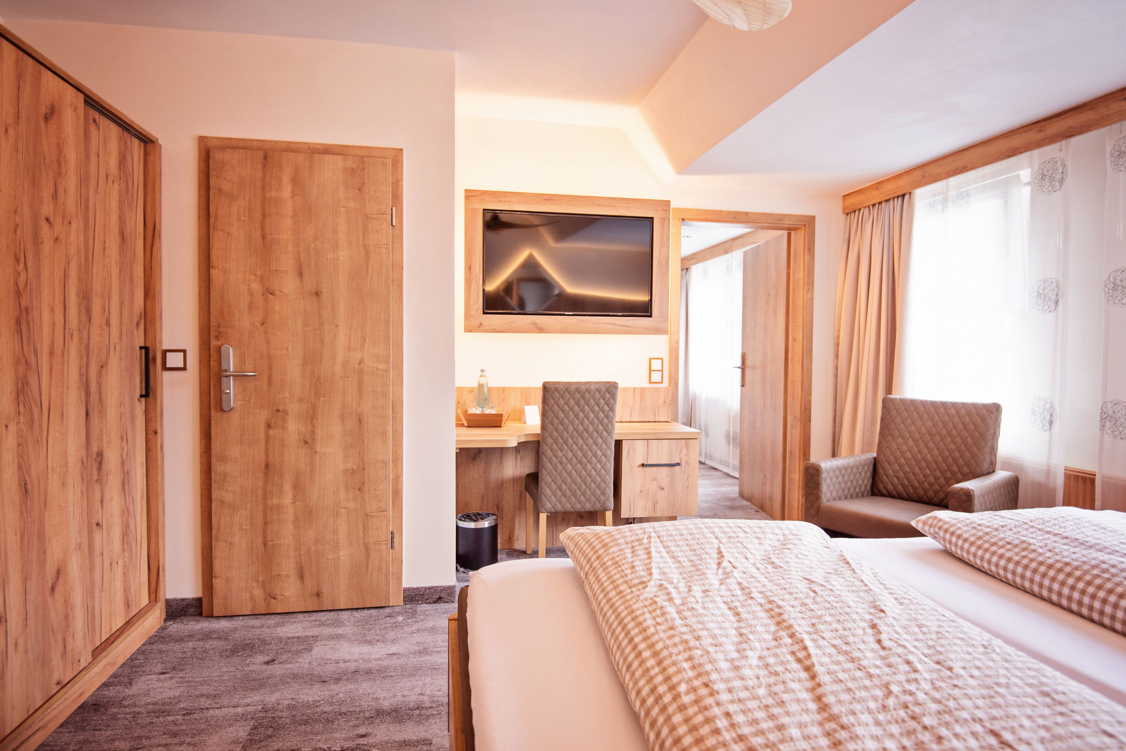 A hotel room with a wooden wardrobe, a desk with a chair, a bed with two pillows, and a large mirror.