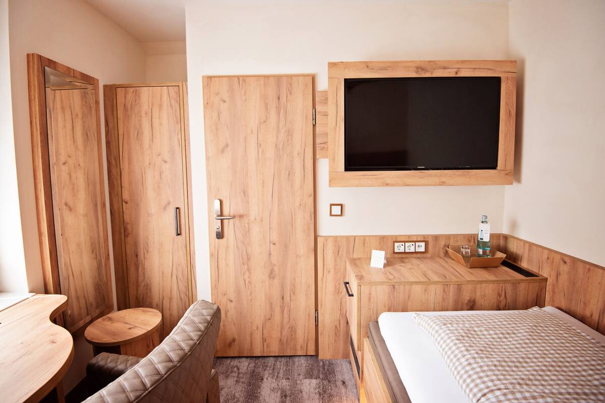 A room with a bed, a wooden desk, a television mounted on the wall, and a wooden wardrobe.
