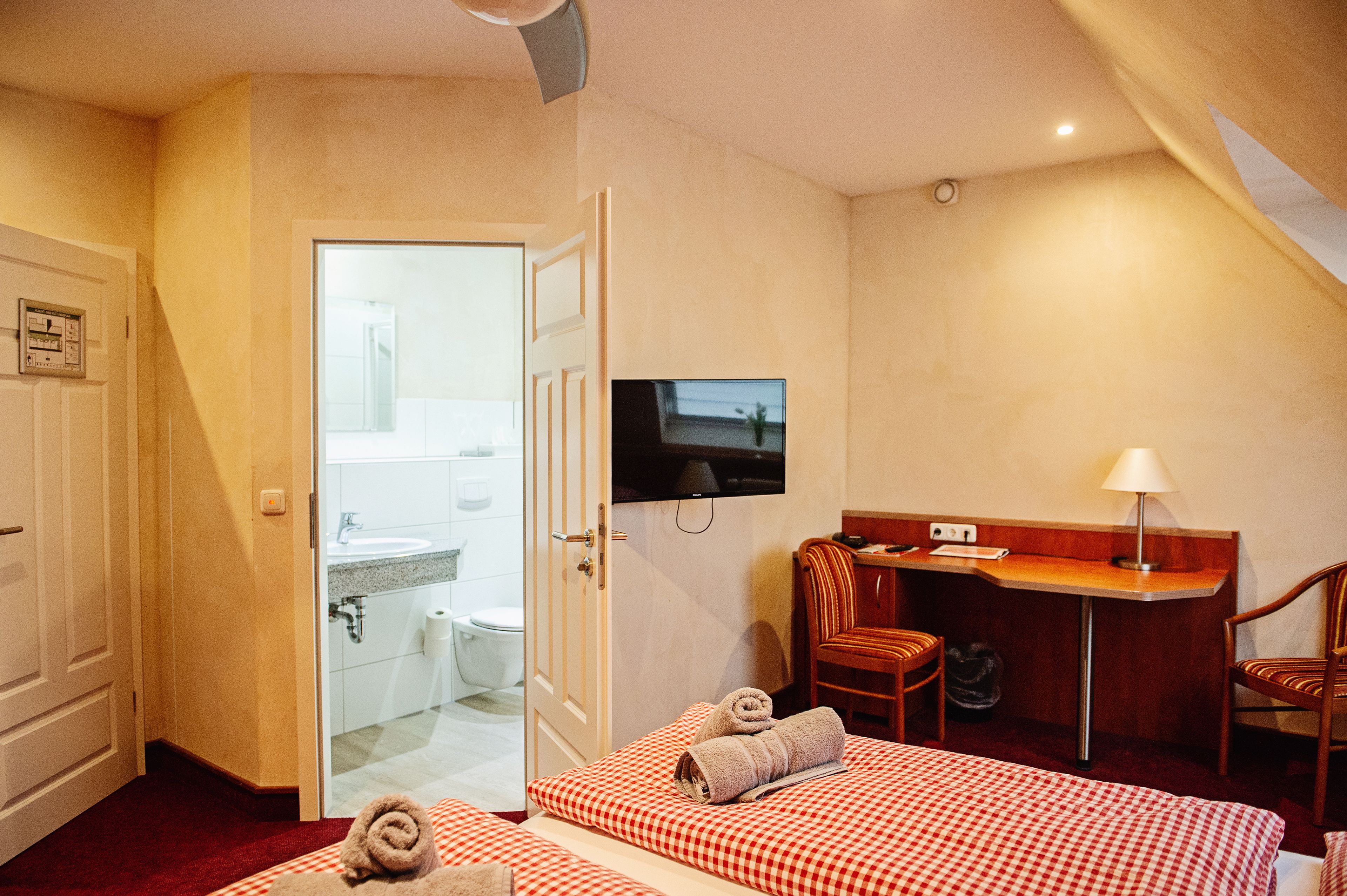 A hotel room with a bed, a desk, a TV, a bathroom, and a red and white checkered bedspread.