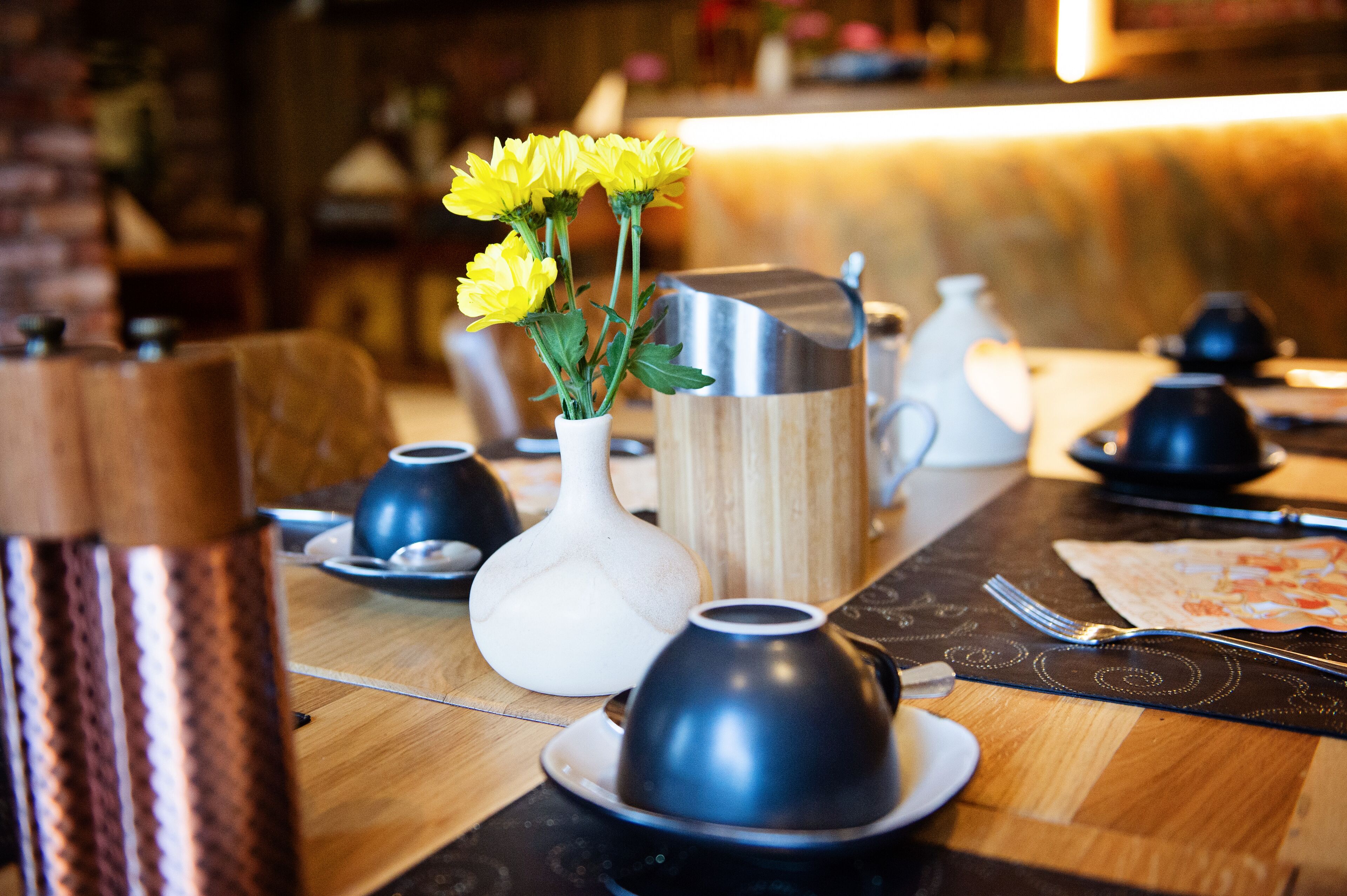 Una mesa de comedor dispuesta con diferentes pots de té y tazas, un florero con flores amarillas y una jarra de madera.