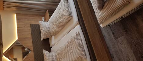 Una habitación con dos camas, cada una con un cojín a cuadros y una toalla enrollada. El piso es de madera y hay una ventana con cortinas de gasa.